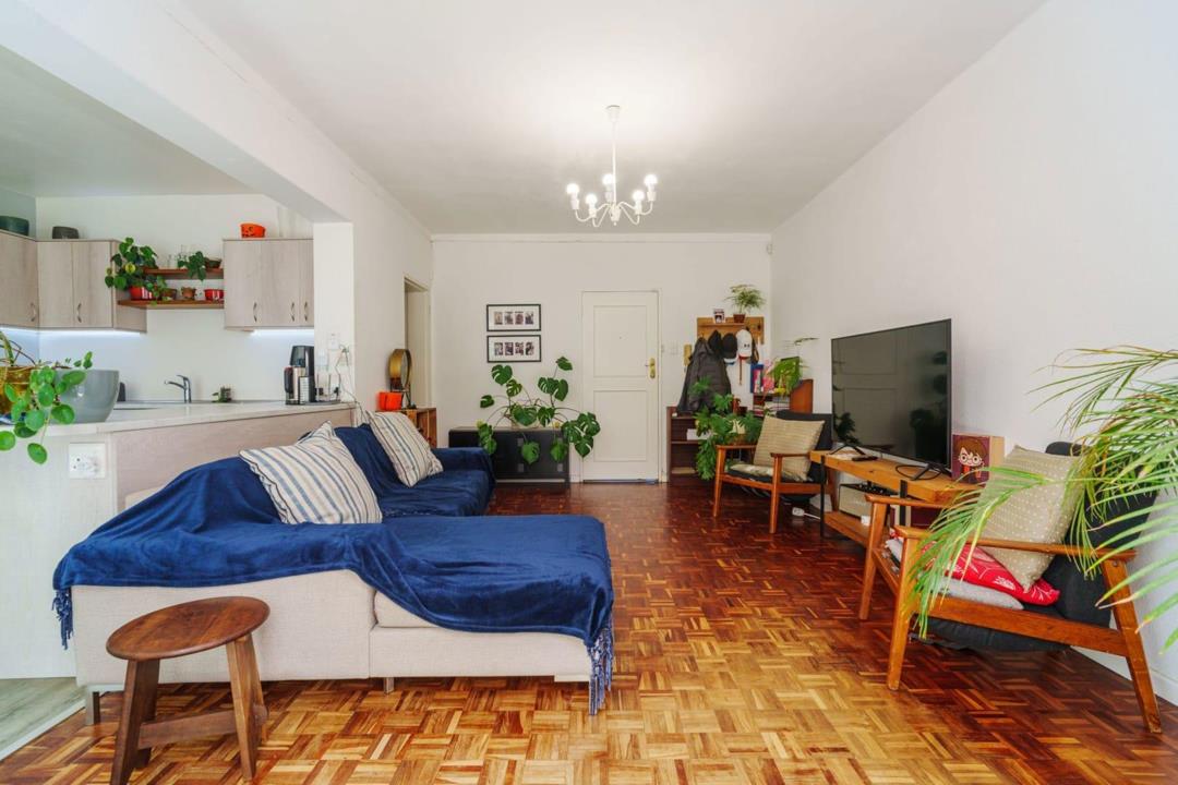 Wide-angle view of the open-plan lounge and kitchen from the garden-facing end, showing the full depth of the living area with parquet floors, blue sofa, mid-century armchairs, flat-screen TV, monstera houseplants, and t