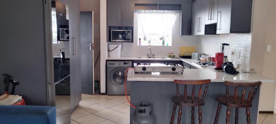 Open-plan kitchen with grey shaker cabinets, white quartz-effect countertop, geometric-tile splashback, double sink, microwave, and a portable two-burner gas hob on the breakfast bar; washing machine visible in adjacent 