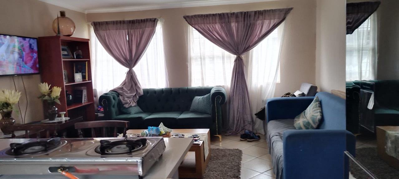 Lounge viewed from the kitchen side showing a large teal Chesterfield sofa, blue armchair, wooden display unit with flat-screen TV, two curtained windows with purple sheer drapes, and a portable gas hob in the foreground