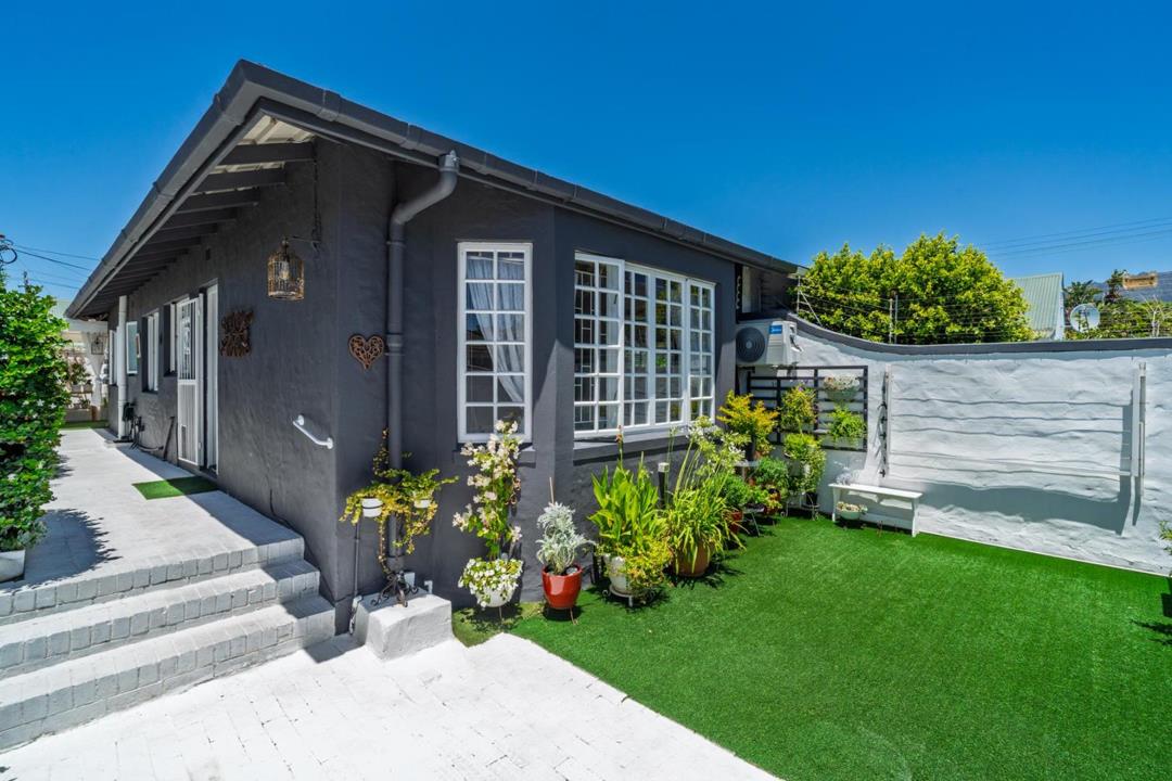 Exterior front-corner view of a single-storey dark grey rendered cottage with white-trimmed cottage-pane windows, a paved driveway with steps, artificial lawn, potted plants, a wall-mounted air-conditioning unit, and a c