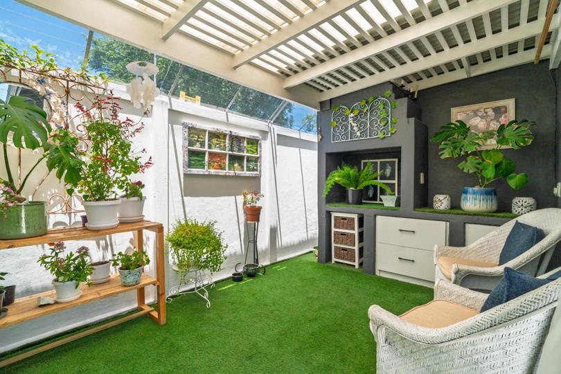 Covered outdoor courtyard entertainment area with a white-painted pergola roof, artificial turf floor, potted plants on a timber shelf, a built-in grey niche unit with white drawers and wicker storage baskets, and two wh