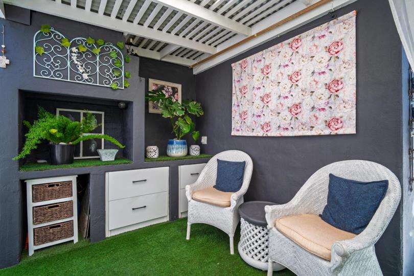 Close-up of the pergola-covered entertainment nook showing two white wicker chairs with navy cushions, a built-in grey recessed shelf displaying ferns and potted plants, white drawer unit with rattan basket storage, and 