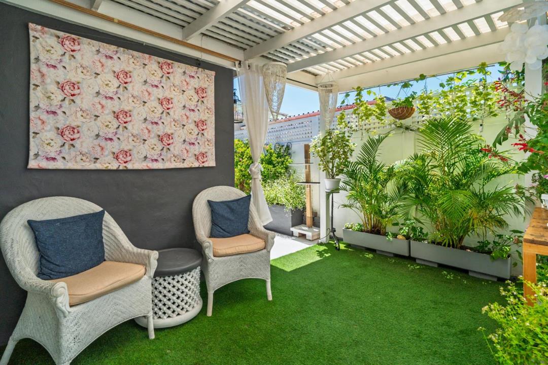 Wide view of the pergola-shaded courtyard seating area with white wicker chairs, a floral wall tapestry, sheer white curtain, bead chandelier, lush tropical potted plants in grey planters along a white boundary wall, and