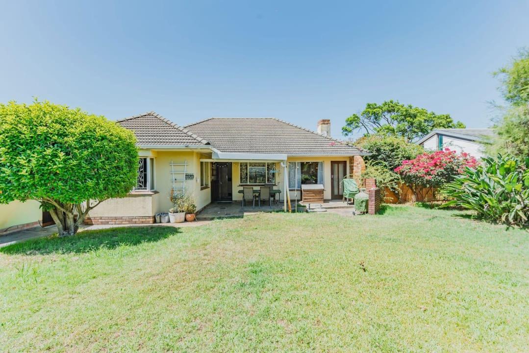 Rear exterior of a single-storey tiled-roof home viewed across a wide, well-kept lawn, showing a covered patio with outdoor furniture, a braai area, bougainvillea, and established trees on a sunny day.
