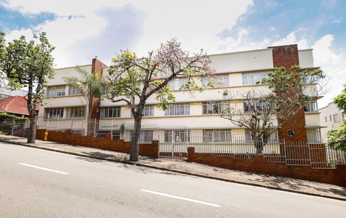 Street-facing exterior of a three-storey mid-century white-rendered apartment block with brick accents, barred windows, mature flowering trees, and a low brick perimeter wall with steel security gates.
