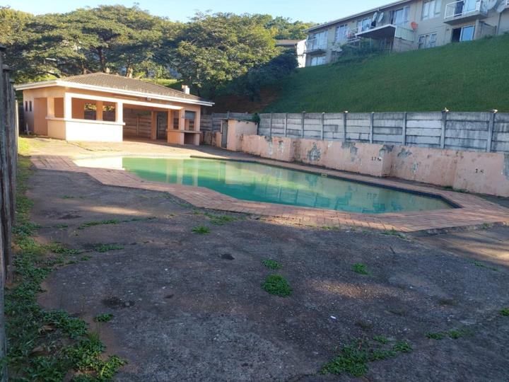 Communal swimming pool area within the complex showing a rectangular pool with green-tinted water, a brick paving surround, an adjacent single-storey entertainment or braai room with a covered patio, and a concrete retai