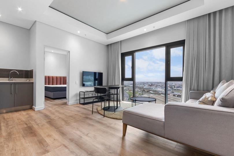 Wide-angle view of the open-plan lounge looking through an open doorway into the bedroom, which reveals a bed with a terracotta upholstered headboard; a partial kitchenette counter and grey cabinetry are visible to the l