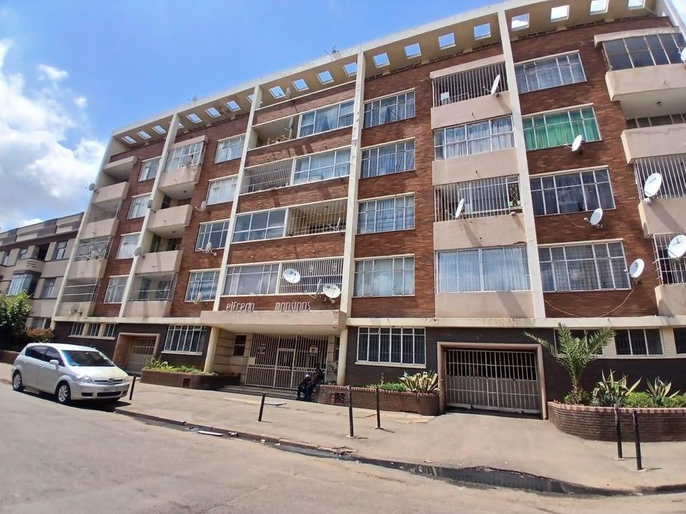Street-level exterior view of Elfrida Mansions, a five-storey mid-century brick apartment building in Yeoville, Johannesburg, showing a gated ground-floor entrance, security-barred windows, multiple balconies, satellite 