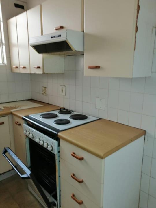 Second angle of the kitchen revealing cream laminate cabinetry, a four-plate white electric stove with extractor hood, laminate countertops, white square wall tiles, and an open oven door with a detached oven rack visibl