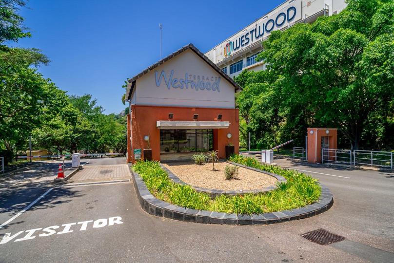 Ground-level view of the manned security gatehouse at the entrance to Westwood Terraces, branded with the complex name and surrounded by landscaped gardens, with Westwood Mall signage visible behind.