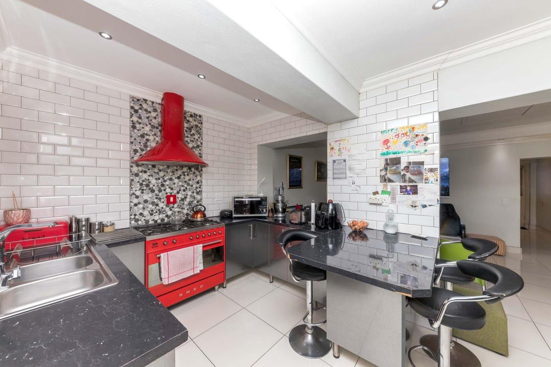 Open-plan kitchen showing a red Smeg range cooker with matching red extractor hood over a black-and-white mosaic splashback, black granite breakfast bar with bar stools, white subway-tiled walls, and recessed downlightin