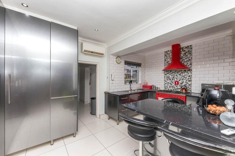Wide-angle kitchen view featuring a tall stainless-steel pantry cupboard on the left, a wall-mounted air-conditioning unit, the red Smeg range cooker in the background, and a curved black granite breakfast island with ba