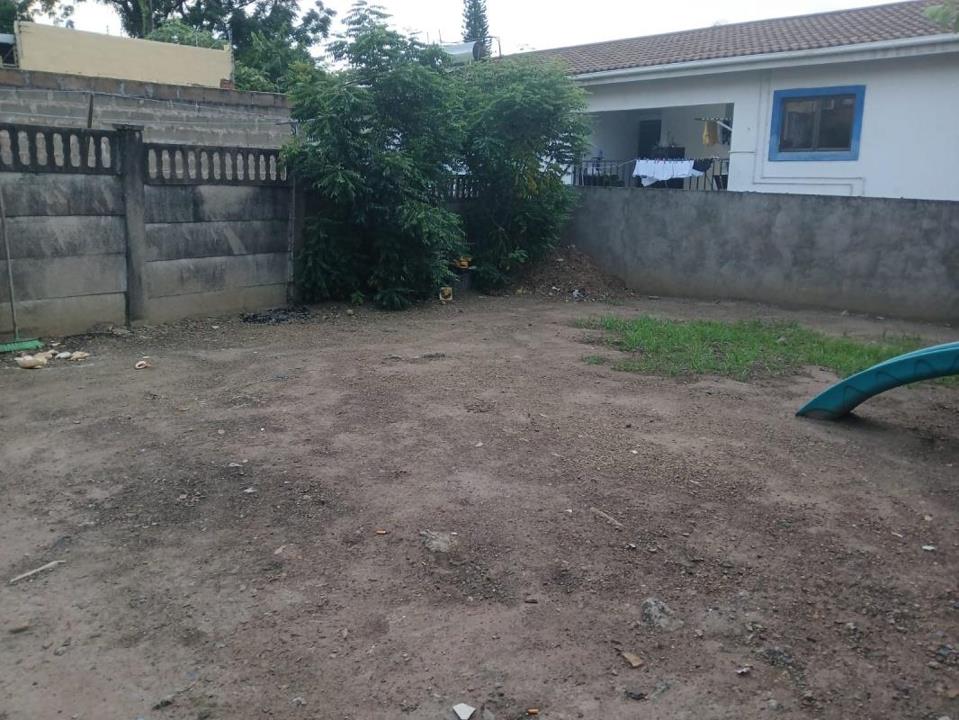 Bare dirt back yard enclosed by concrete block walls, with a mango or similar fruit tree in the far corner, patchy grass, a children's plastic slide visible at the right edge, and a neighbouring single-storey white house