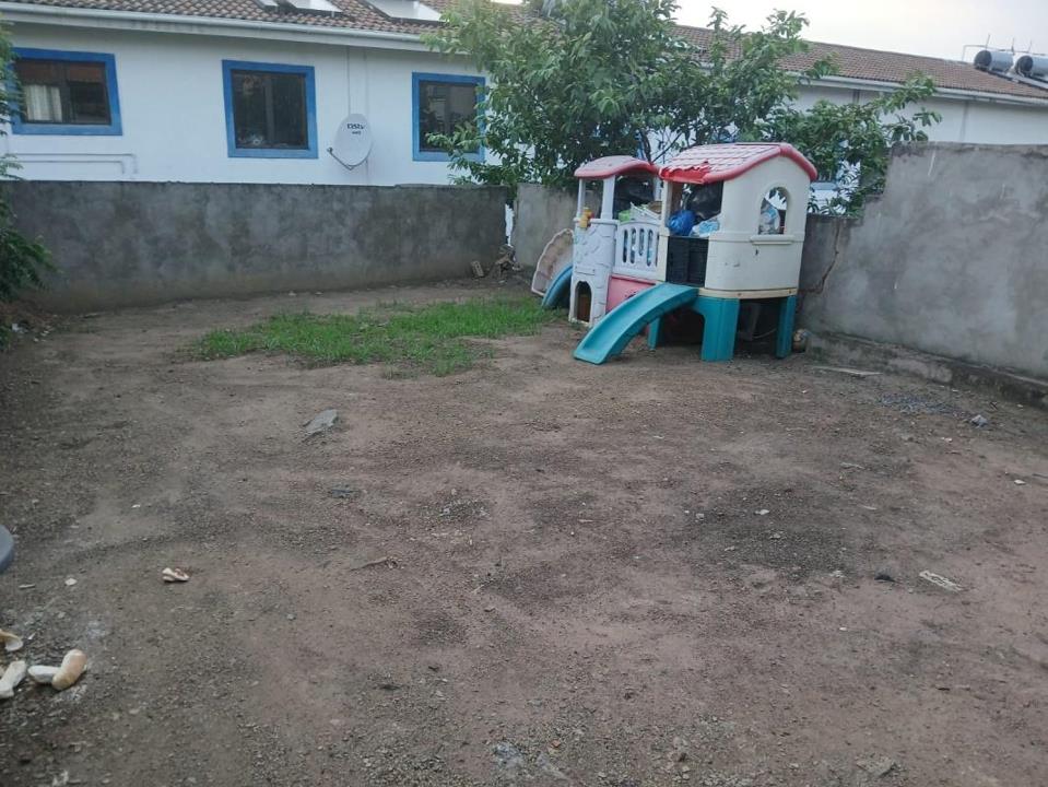 Back yard showing a plastic children's playhouse with a teal slide set against a concrete boundary wall, a DStv satellite dish on the neighbouring white house, a tree, and a dirt surface with sparse grass patches.