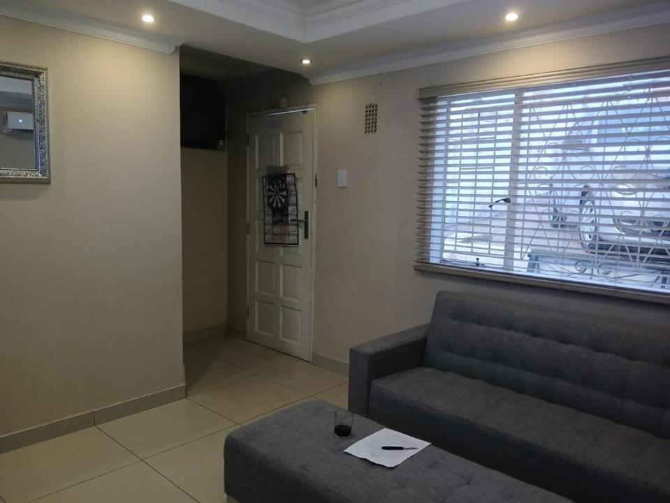 Interior lounge/living room with large grey fabric sofa, tiled flooring, recessed downlighting set into a coved ceiling with cornicing, a white panel front door with a dart board mounted on it, venetian blinds on a secur