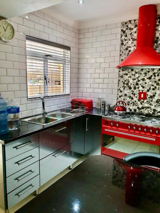 Kitchen corner view showing a stainless-steel double sink beneath a window with horizontal blinds, gloss-grey base cabinets, black granite countertop, and the red Smeg range cooker with matching hood to the right.