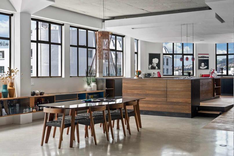 Open-plan kitchen and dining area with a large timber-veneer island, integrated gas hob, an eight-seat dining table with timber chairs under a sculptural copper pendant light, warehouse-style multi-pane steel windows, an
