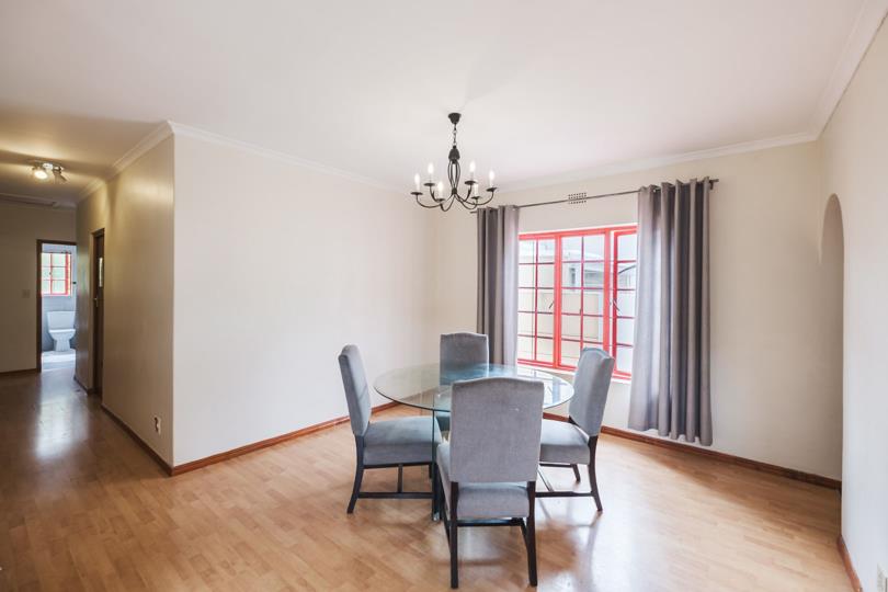 Formal dining room featuring a round glass-top table surrounded by five grey upholstered chairs beneath a black wrought-iron candelabra chandelier, with timber laminate flooring, white walls with crown moulding, and a re