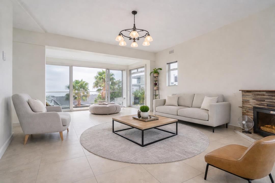 Spacious lounge with large-format tile flooring, a light-grey three-seater sofa and mid-century style armchairs arranged around a square wooden coffee table on a round rug, a stone-clad electric fireplace to the right, a