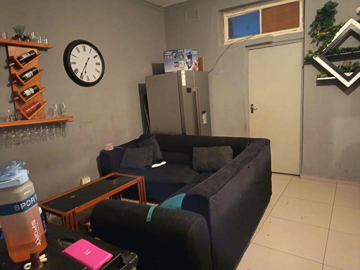 Alternative angle of the living area showing the wall-mounted wine rack, stemware shelf and Roman-numeral wall clock on grey-painted walls, a double navy sofa and glass-top coffee table, the stainless-steel fridge-freeze