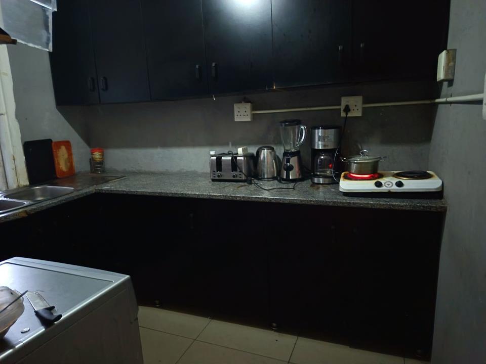 Second view of the same kitchen showing the full L-shaped dark laminate cabinetry run, granite-effect worktop, toaster, kettle, blender, coffee maker, and portable electric hotplate, with a stainless-steel sink at the le