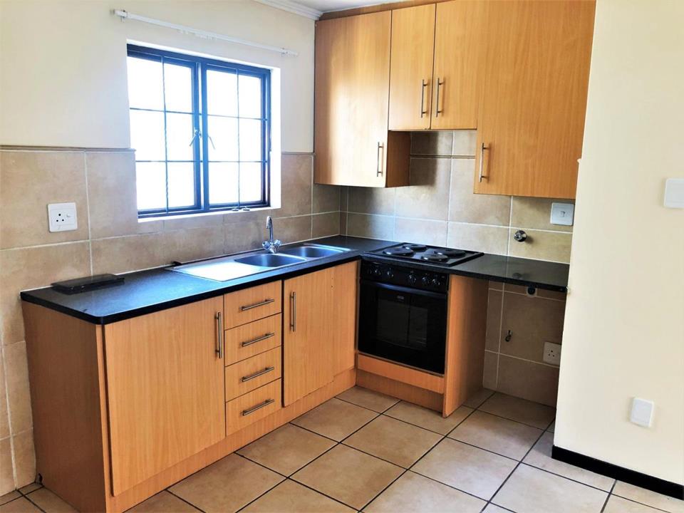 L-shaped kitchen with oak-finish base and wall cabinets, dark granite countertops, double stainless-steel sink under a security-barred window, and a black freestanding stove; beige ceramic floor tiles and tile splashback