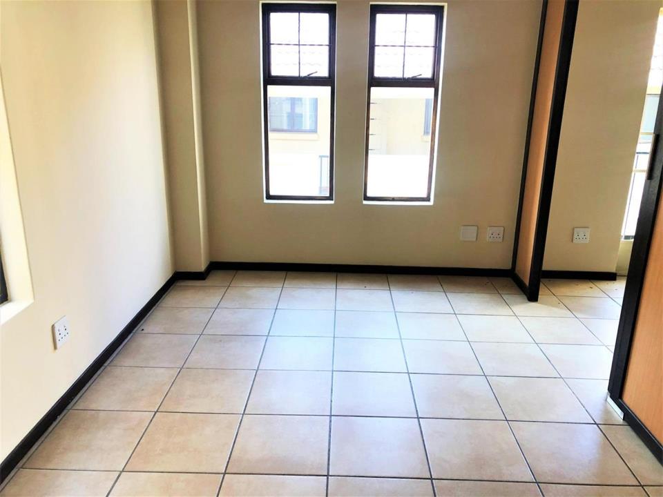 Empty upper-level loft room or second bedroom with beige ceramic tiles, two double-hung windows with security grilles overlooking the complex, multiple double power outlets, and a doorway on the right.