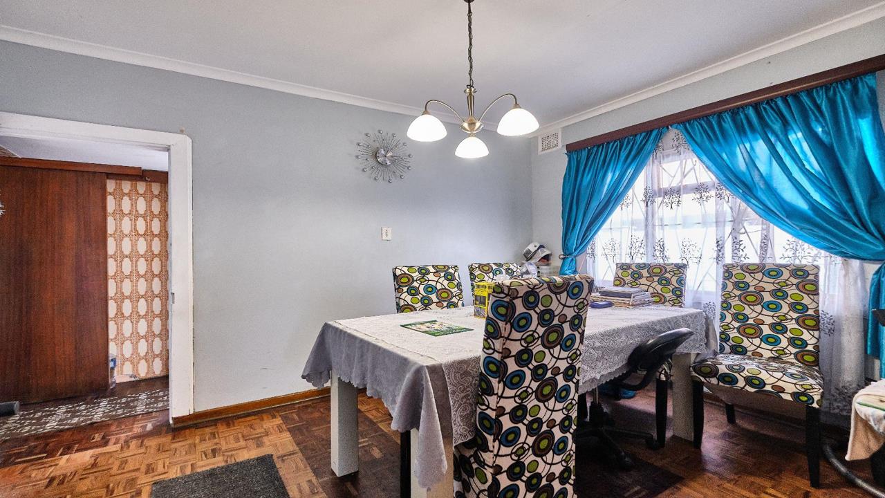 Dining room with original parquet flooring, a six-seater dining table with patterned chair covers, a brass-finish chandelier, white corniced ceiling, turquoise curtains with sheer lining over a large window, and a retro 