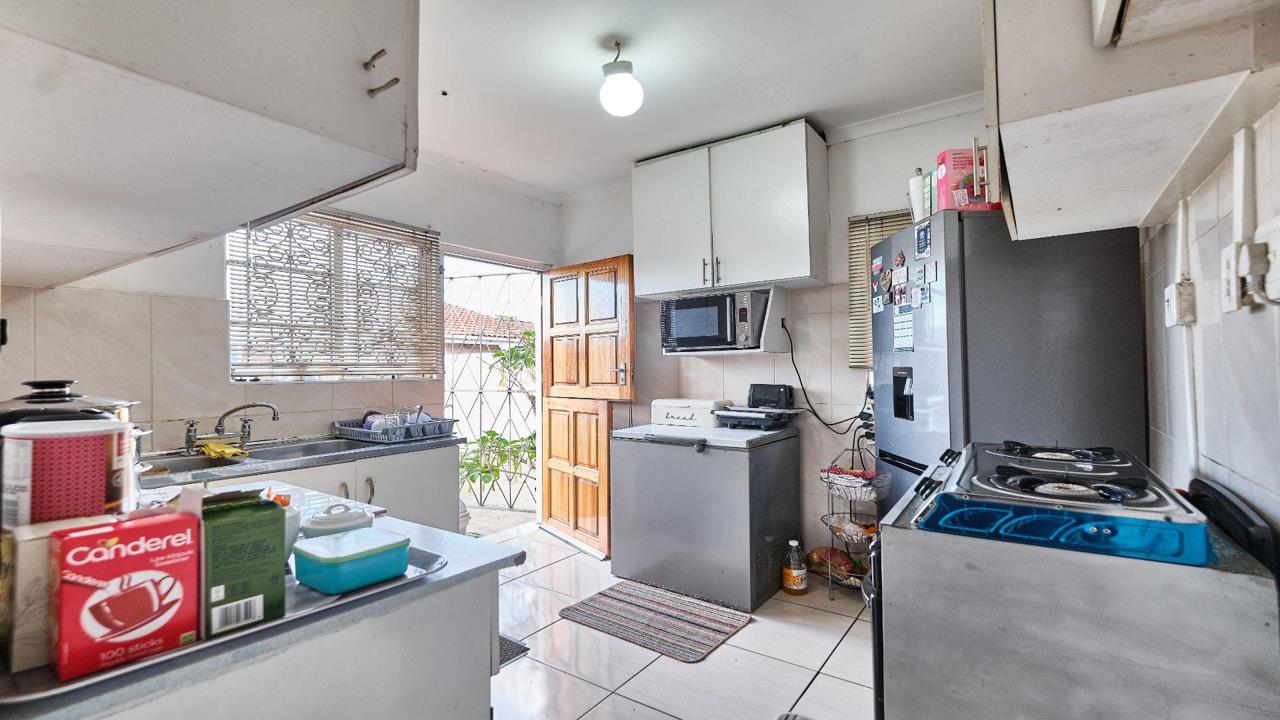 Larger kitchen viewed from the doorway showing a U-shaped layout with white cabinets, a double sink, a chest freezer, a microwave, a stainless-steel fridge, an electric hob, tiled floors, and a wooden security door open 