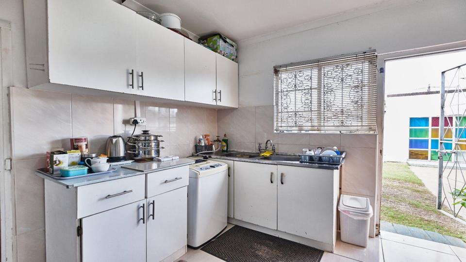 Compact fitted kitchen with white upper and lower cabinets, tiled splashback, a dark countertop sink beneath a burglar-bar window with venetian blinds, a washing machine integrated under the counter, and an open wooden s