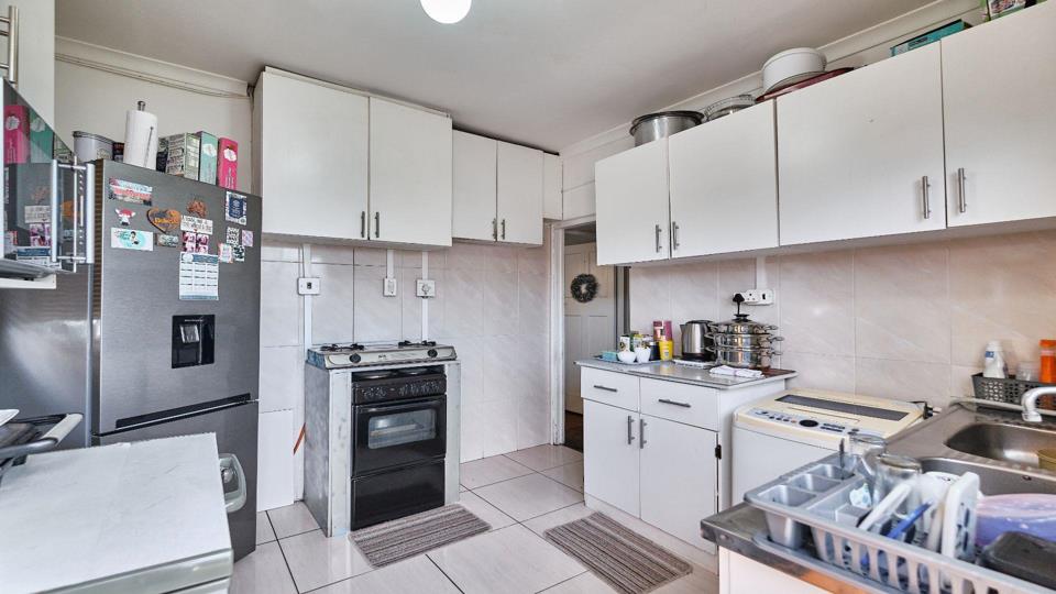 Second angle of a fitted kitchen showing white upper and lower cabinets, a freestanding gas stove with oven, a stainless-steel fridge-freezer with water dispenser, tiled splashback, a washing machine, and tiled flooring.