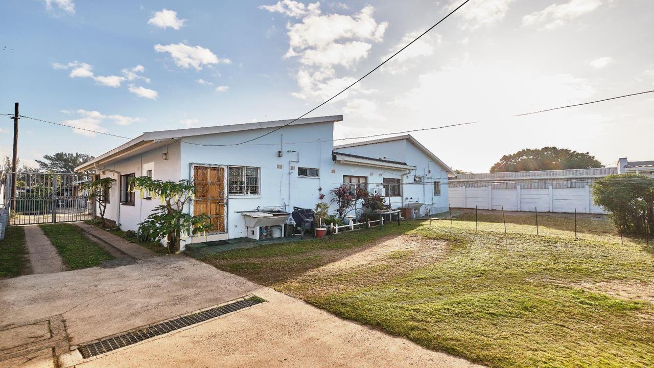 Exterior front view of a white-painted semi-detached house in Merewent, Durban, showing a flat and pitched metal roof, burglar-bar-fitted windows and a wooden security door, a small garden with potted plants, a concrete 