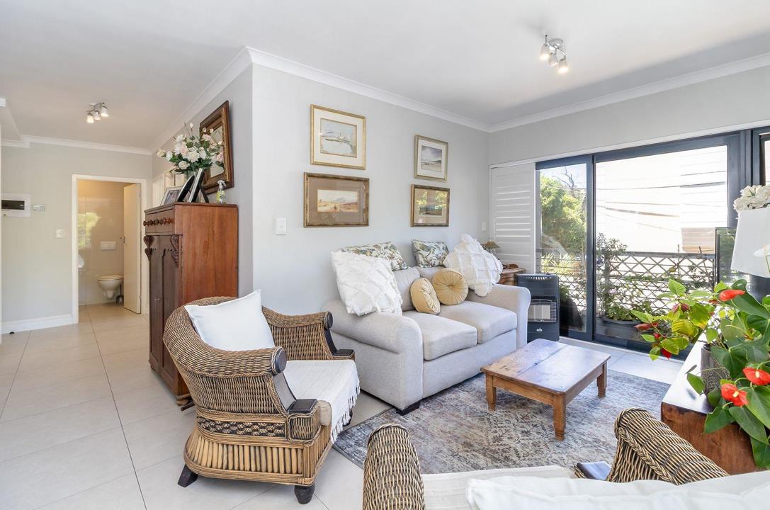 Bright open-plan lounge with grey upholstered sofa, rattan armchairs, rustic wooden coffee table, framed landscape artwork on grey walls, large-format white floor tiles, and a sliding glass door opening to a balcony with