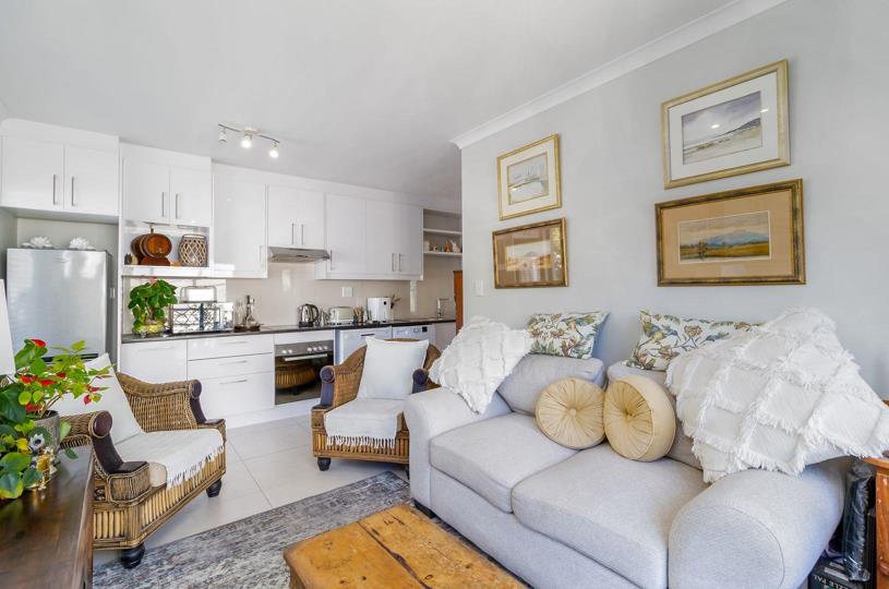 Open-plan view showing the white gloss kitchen with dark countertops and under-counter oven flowing into the lounge area featuring rattan chairs, grey sofa, and framed artwork, all under crown moulding on grey-white wall