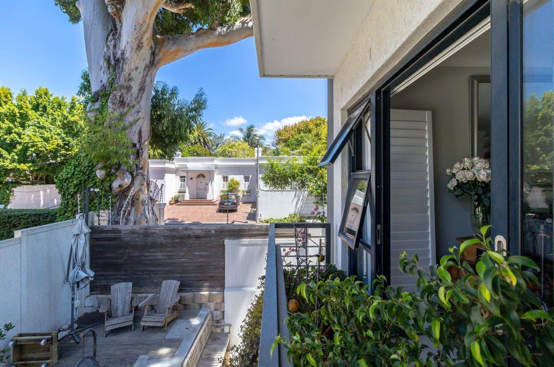 Private timber-decked balcony with Adirondack chairs, established eucalyptus tree canopy overhead, lush surrounding greenery, and a view towards a neighbouring white Cape Dutch-style house under blue skies.