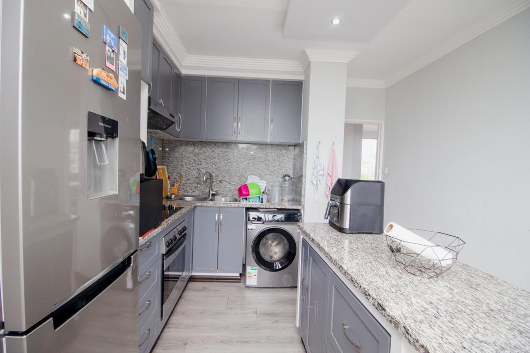 Galley-style kitchen viewed from the entrance showing grey shaker cabinets, granite-look countertops, stainless-steel fridge-freezer with water dispenser, front-loader washing machine, air fryer on the counter, and light