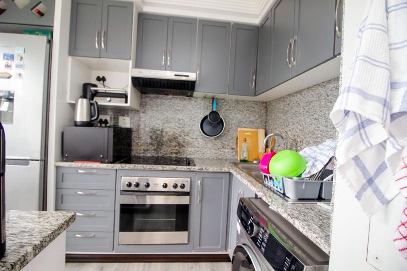 Kitchen angle showing stainless-steel oven and hob beneath an extractor fan, grey upper and lower cabinets with granite-look splashback, microwave and kettle on the counter, and front-loader washing machine to the right.
