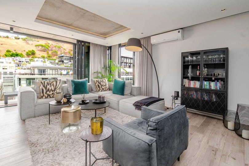 Living room showing the grey L-shaped sectional sofa, velvet accent armchair, round coffee tables with gold accents, a shag rug, wall-mounted split air-conditioning unit, a black glass-door display cabinet, and Signal Hi