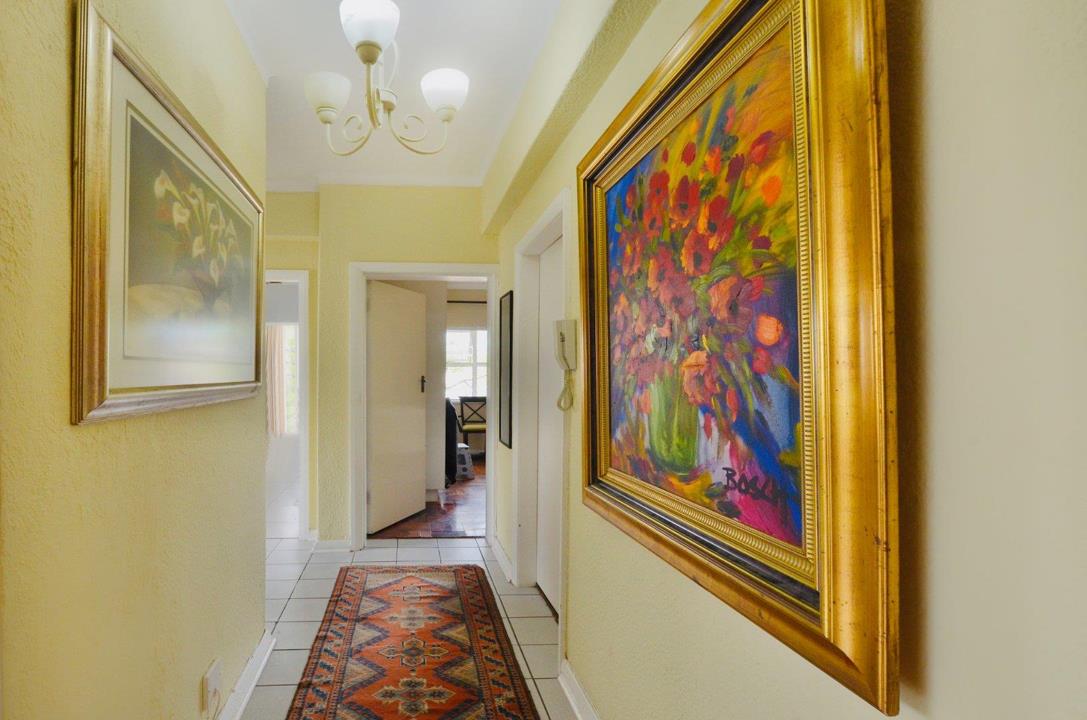 Tiled entrance hallway with cream walls, a decorative three-arm chandelier, intercom handset on the wall, Persian-style runner rug, and two framed oil paintings including a large floral canvas in a gold frame.