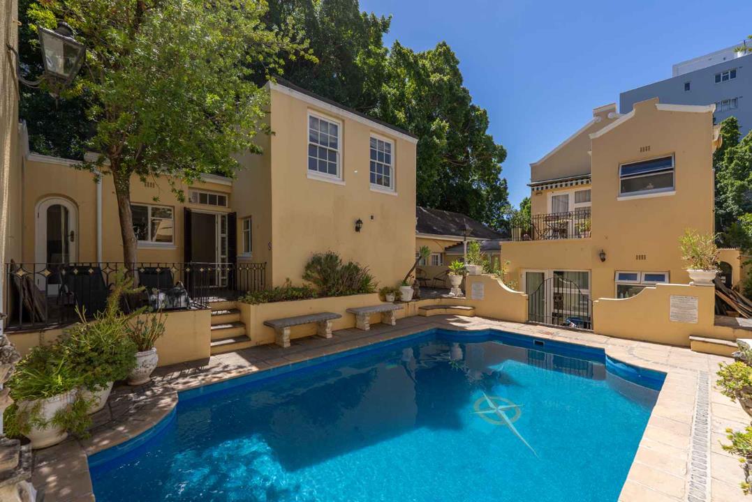 Rear exterior of a cream-rendered double-storey heritage townhouse complex showing a sparkling blue communal pool surrounded by a tiled terrace, stone benches, potted plants, and wrought-iron balustrades under mature can