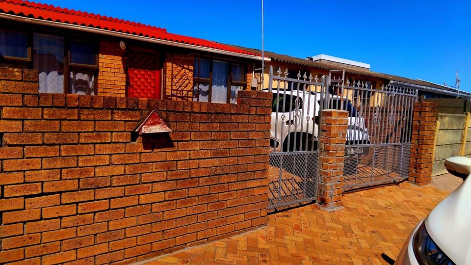 Street-facing exterior of a single-storey face-brick house under a clear blue sky, showing a red corrugated-tile roof, a full-height brick boundary wall with a triangular letterbox, and an ornamental silver steel sliding