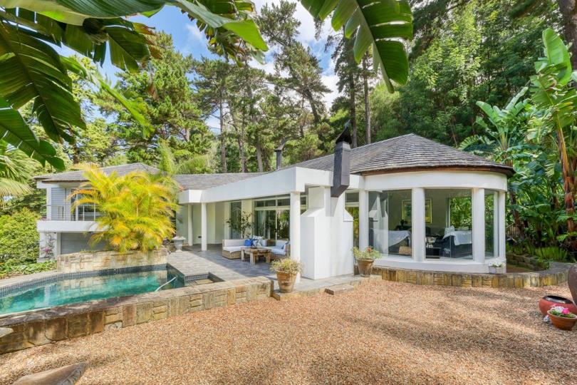 Wide exterior shot of the multi-level white-render home showing the curved rotunda wing with columns and full-height glazing, the covered patio with pool in the foreground, a gravel courtyard, and surrounding pine and tr