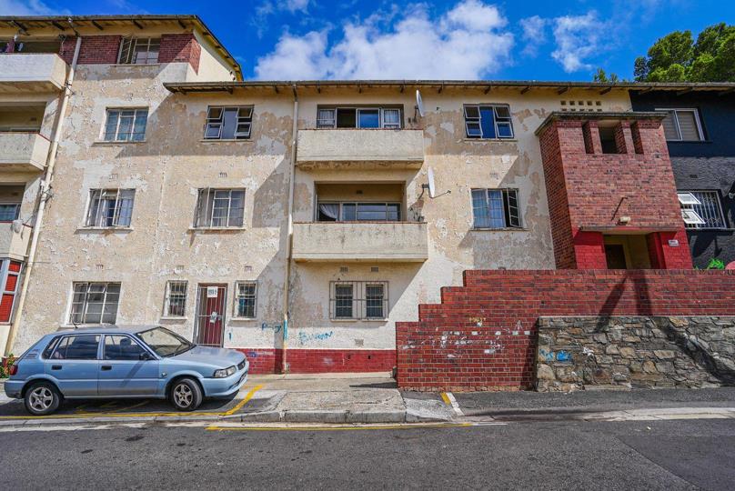 Street-level exterior shot of the Schotschekloof Flats building showing a three-storey rendered facade with a red-and-white base, security bars on windows, a small balcony, and a light-blue hatchback parked in front.