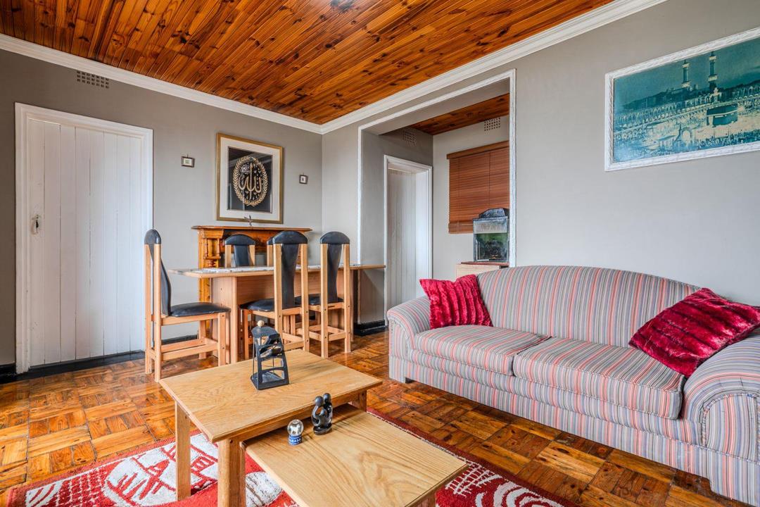Living and dining area with warm tongue-and-groove timber ceiling, parquet wood block flooring, a striped fabric sofa, wooden dining table with four high-back chairs, and a doorway leading to an adjacent room.