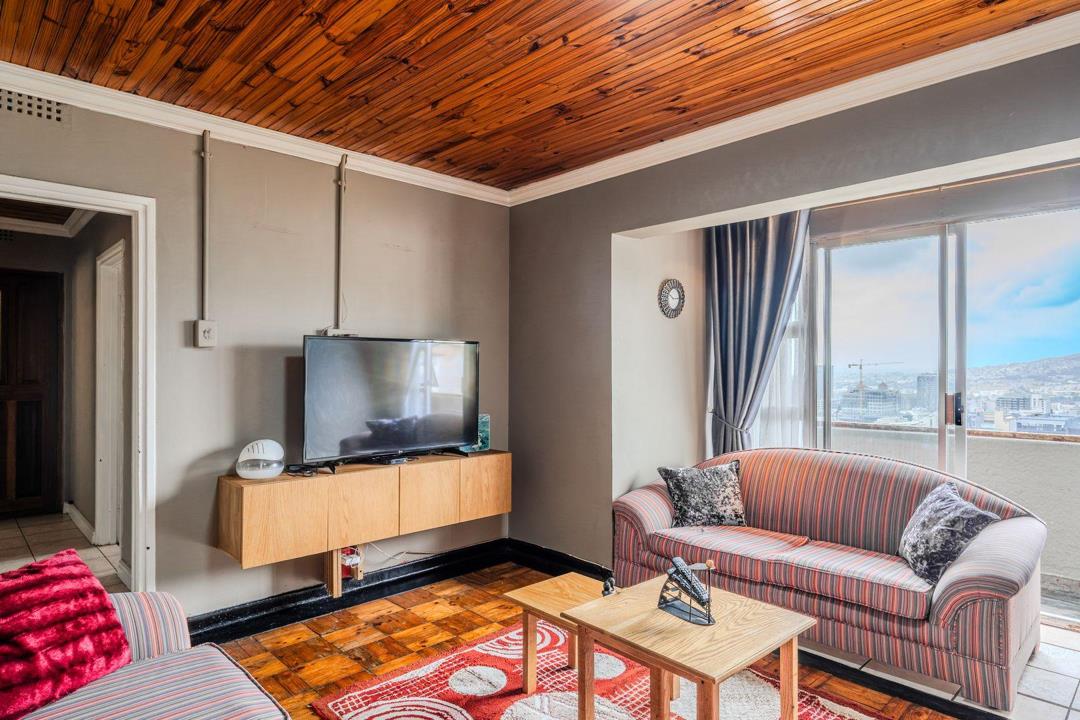 Lounge area with tongue-and-groove timber ceiling, parquet flooring, wall-mounted flat-screen TV on a floating wooden unit, two striped sofas, and a large sliding glass door with city-skyline views beyond.