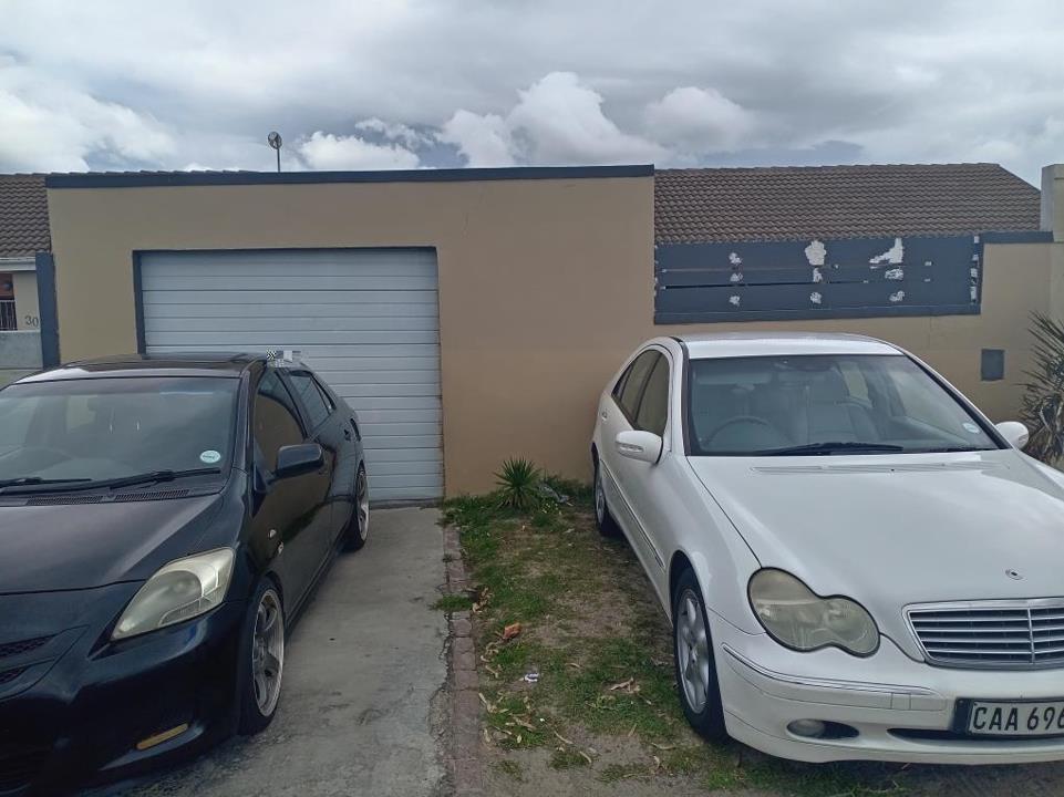 Front exterior of a single-storey house in Pelican Park showing a tan and dark-grey rendered façade, a white sectional roller garage door, a dark-grey slatted boundary wall with partially peeled panel signage, and two ca
