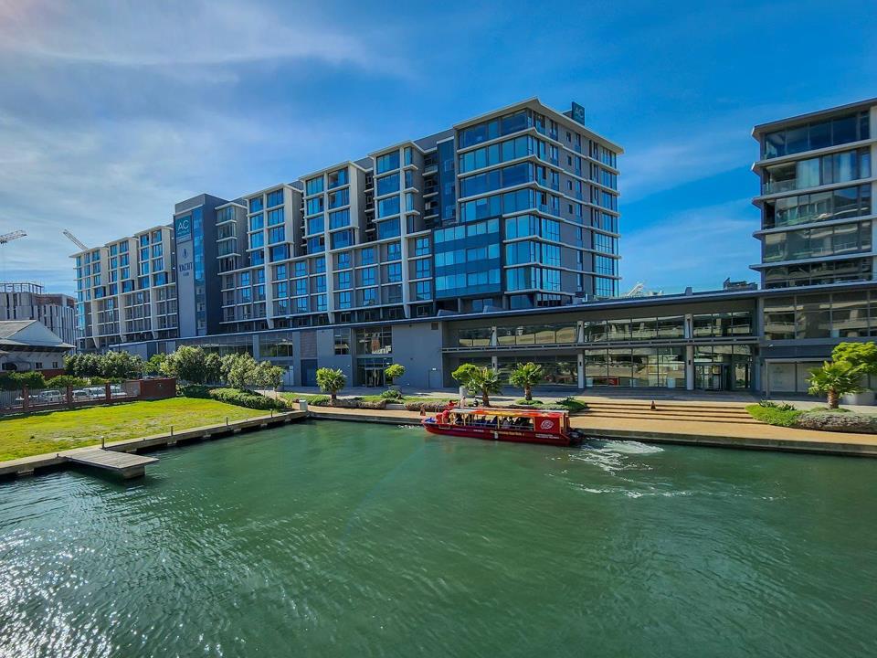 Aerial canal-level view of the Yacht Club development on the Cape Town Foreshore, showing the multi-storey glass-and-concrete residential towers fronting a green waterway, with a red tour boat passing the landscaped prom