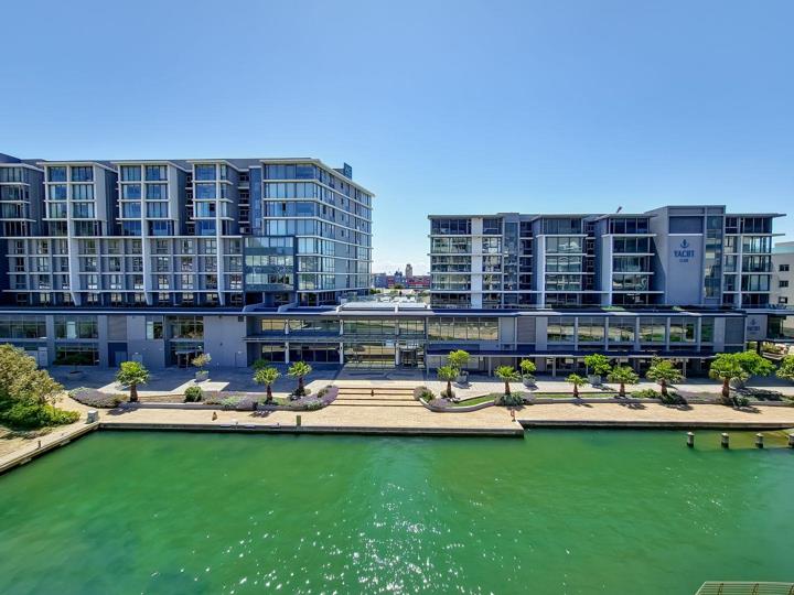 Elevated daytime view of the Yacht Club residential complex from across the marina, showing two glass-facade towers flanking a retail podium level, with a paved waterfront promenade lined with palms and ornamental trees.
