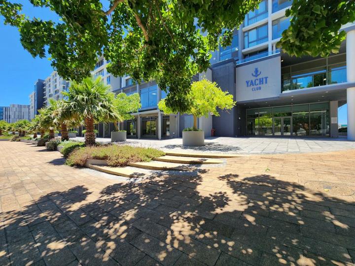 Street-level entrance view of the Yacht Club building lobby with the 'Yacht Club' anchor-logo signage above glass double doors, flanked by mature trees, potted palms, and a paved cobblestone forecourt.