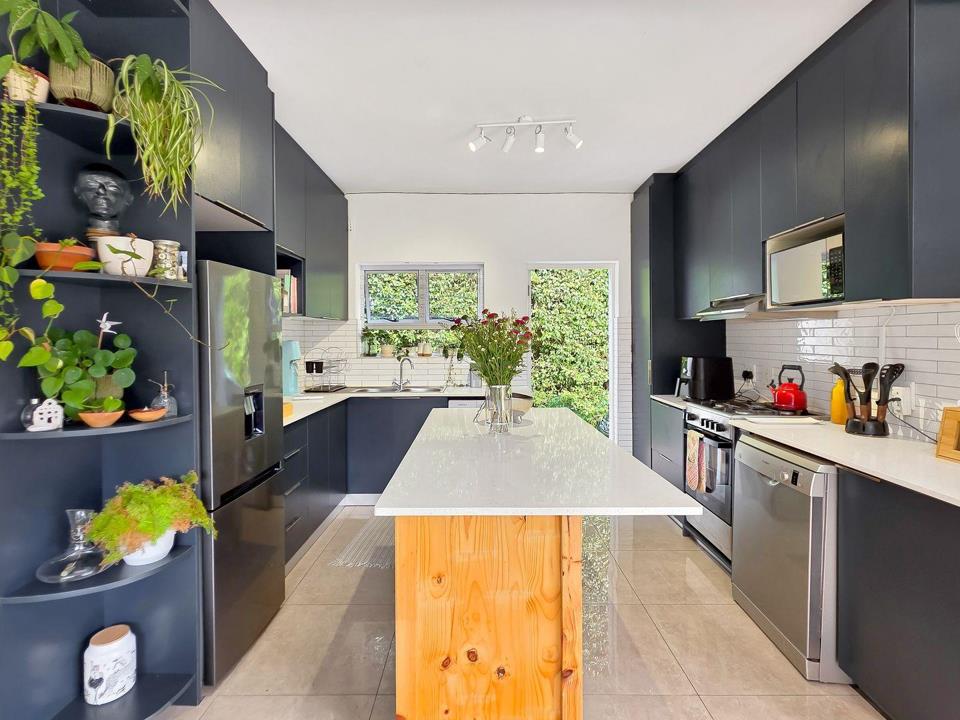 Galley-style kitchen viewed from the island end, showing full-height navy matte cabinetry on both sides, white engineered-stone island with pine timber base, white subway-tile splashback, stainless-steel appliances, and 